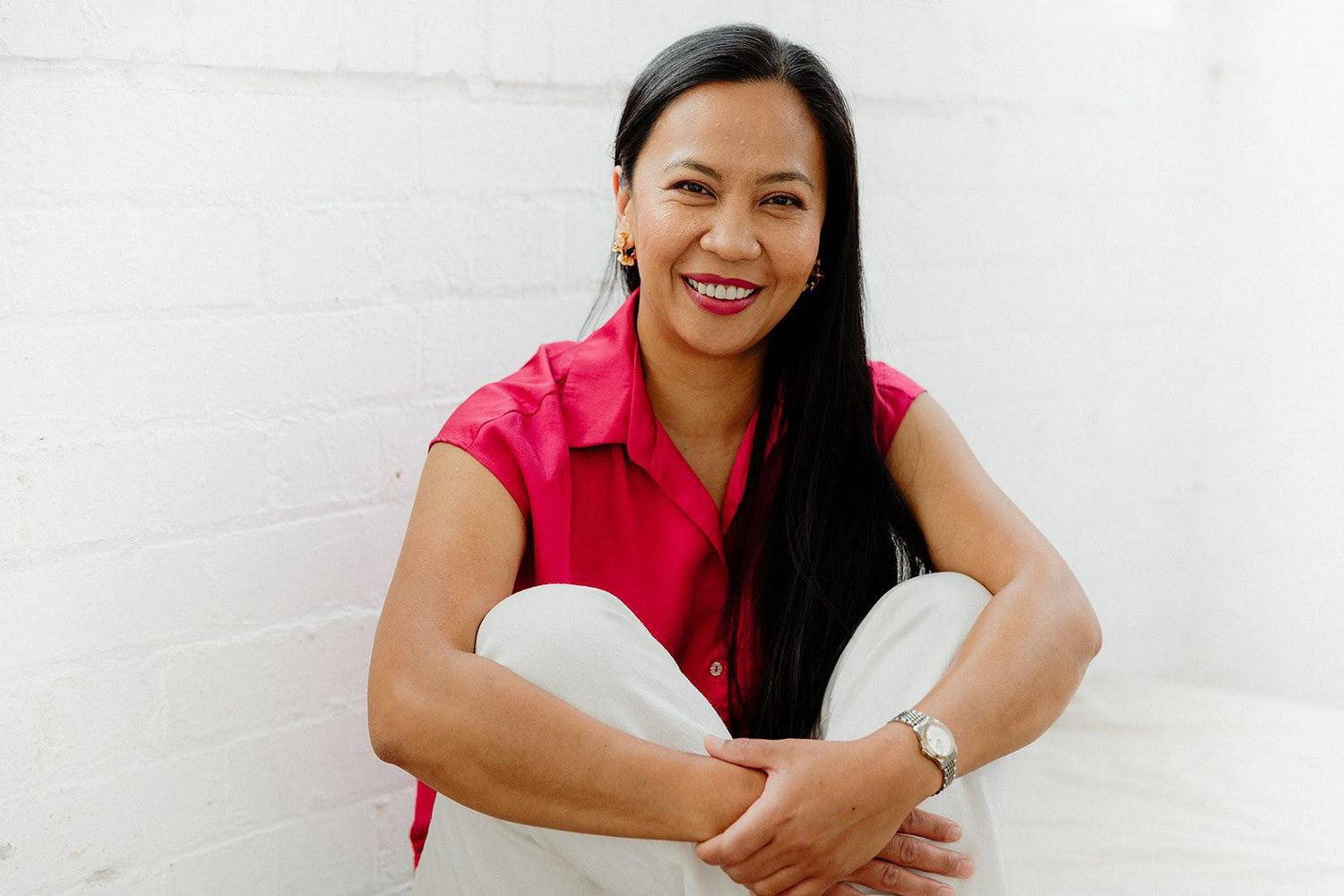 A smiling woman with long black hair sits cross-legged against a white brick wall, wearing a bright pink short-sleeve button-up shirt and white pants.