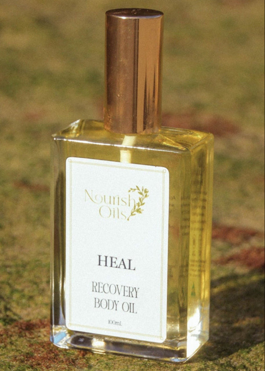 A bottle of Nourish Oils 'Heal' Recovery Body Oil, filled with golden-yellow oil and topped with a metallic gold cap, resting on a grassy ground.