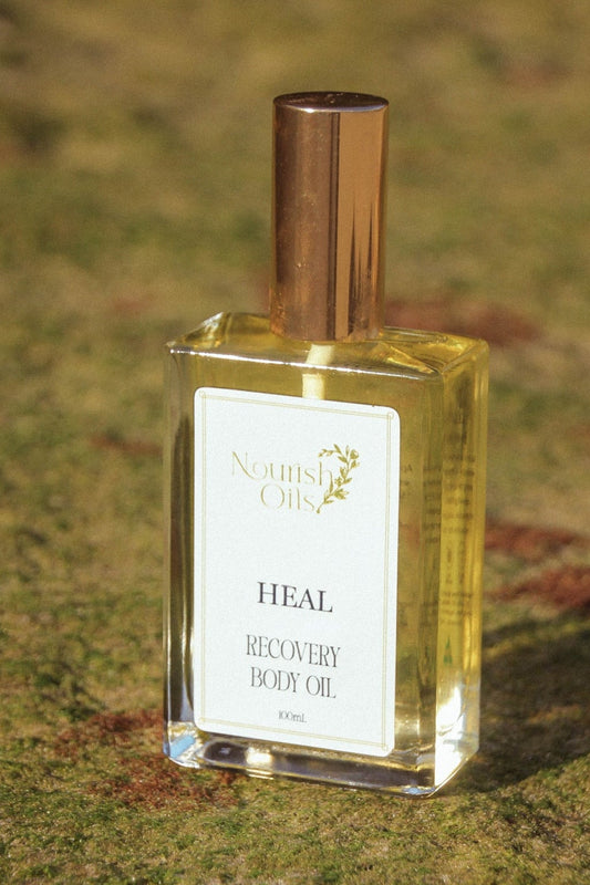 A bottle of Nourish Oils 'Heal' Recovery Body Oil, filled with golden-yellow oil and topped with a metallic gold cap, resting on a grassy ground.