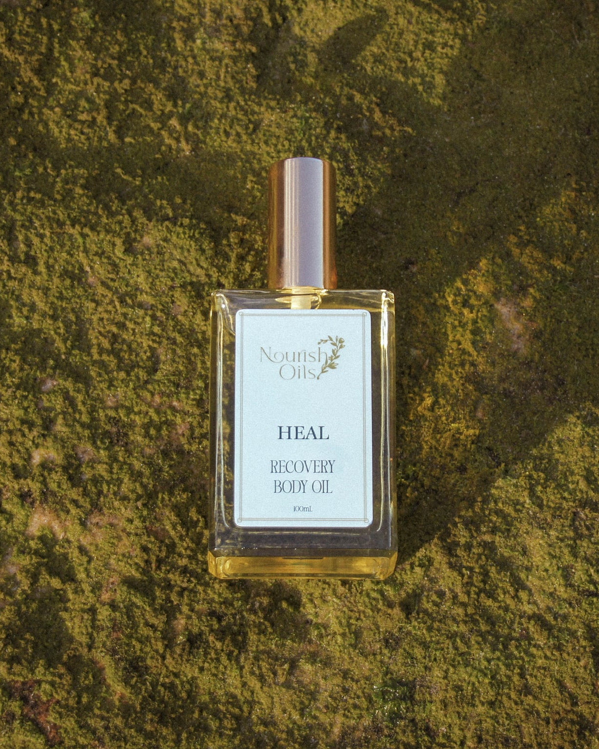 A bottle of Nature's Cures Heal Balm Balmy Oil with a white label and metallic cap, centered on a textured green moss background.