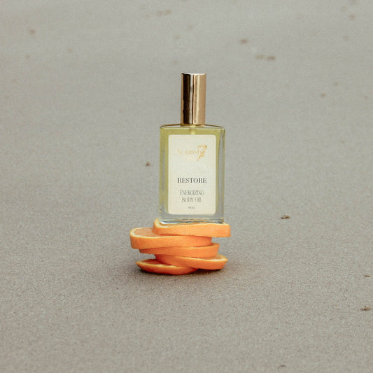 A small rectangular perfume bottle with a gold cap stands on a spiral of orange peel slices against a neutral gray background. The bottle label reads 'RESTORE' and 'FRESH BREEZE'.
