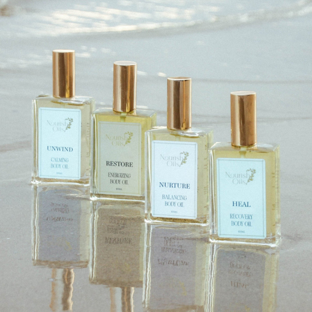 Four small perfume bottles with pale blue labels and metallic gold caps are arranged on wet sand at the beach, with soft water reflections below.