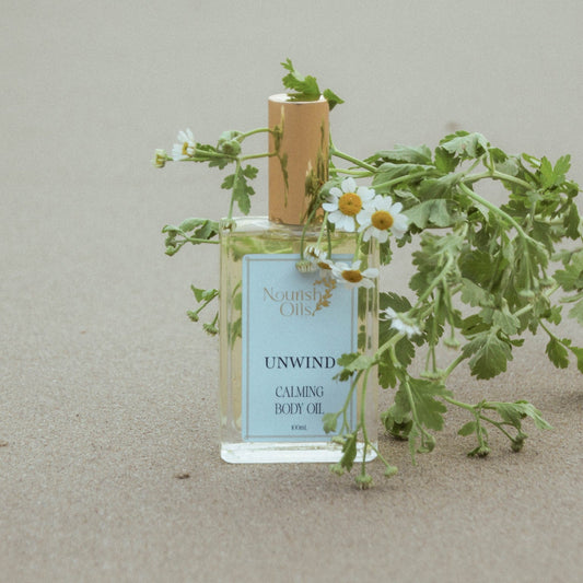 A bottle of Nourish Chose Unwind Calming Body Oil styled with fresh green herbs and white chamomile flowers on a neutral beige background.