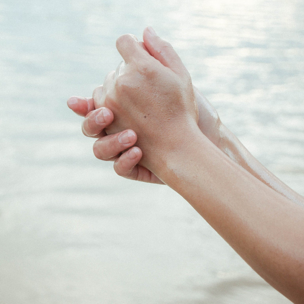 Two hands gently clasped together in front of a softly blurred body of water under bright, hazy sunlight.