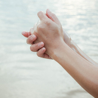 Two hands gently clasped together in front of a softly blurred body of water under bright, hazy sunlight.