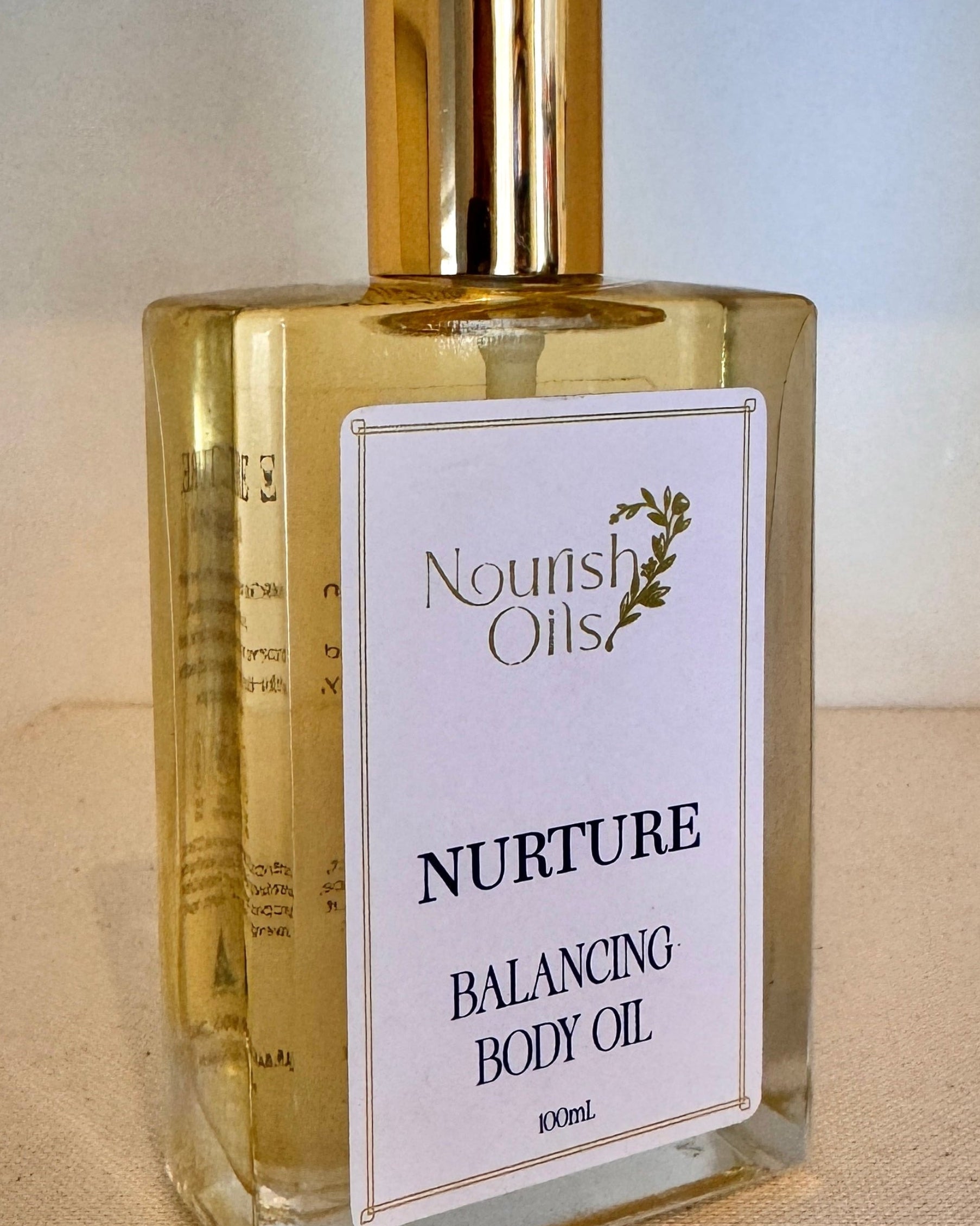 Bottle of Nourish Oils Nurture Balancing Body Oil on a beige surface with a blurred background