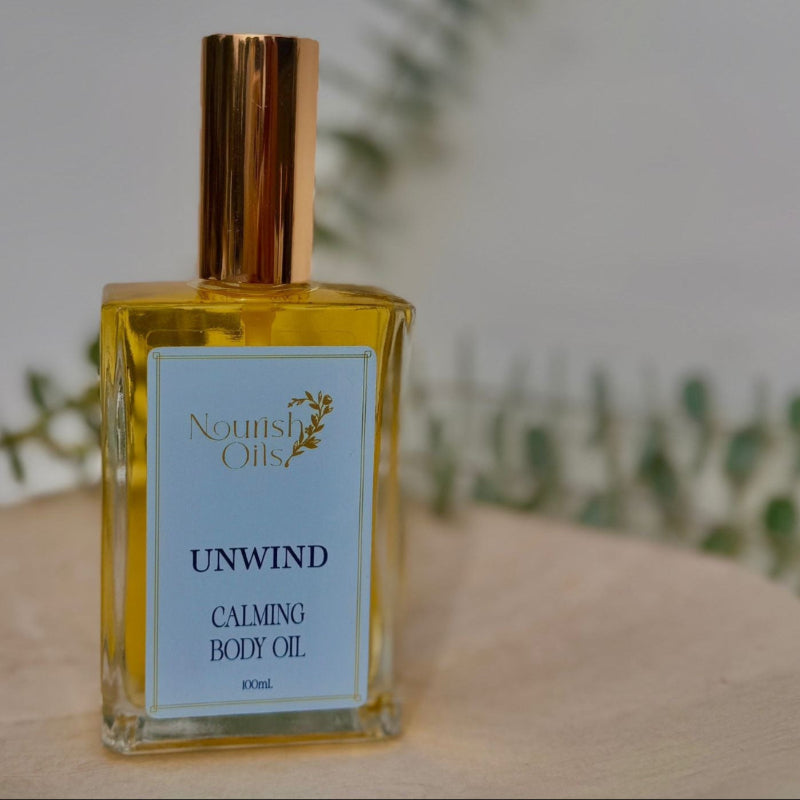 Bottle of Nourish Oils Unwind Calming Body Oil on a wooden surface with a blurred green plant background.
