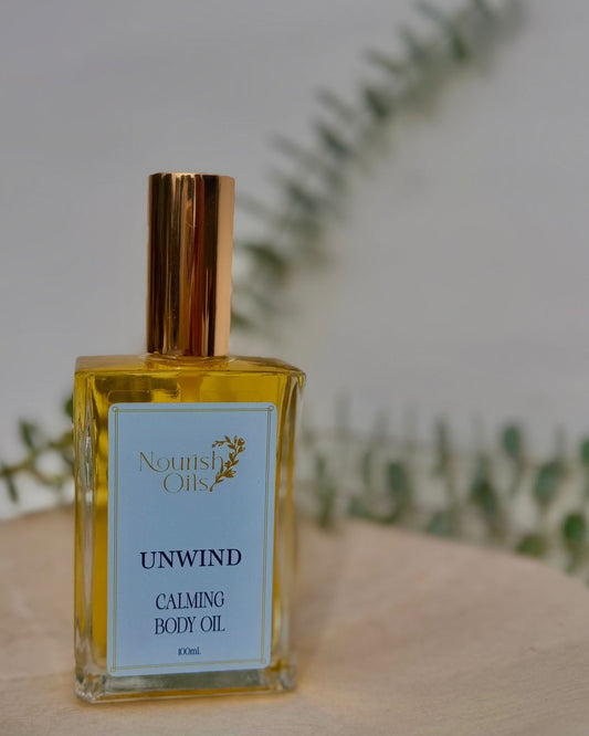 Bottle of Nourish Oils Unwind Calming Body Oil on a wooden surface with a blurred green plant background.