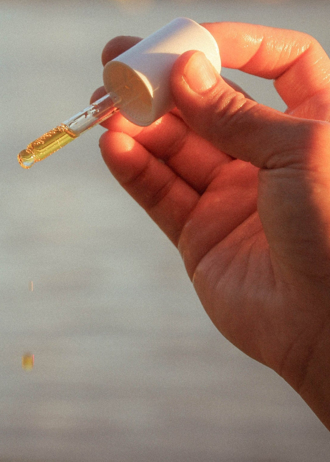 A hand holding the cap off a small white dropper bottle, with a single drop of golden-yellow liquid dripping below. The background is a soft, out-of-focus natural outdoor scene in warm light.