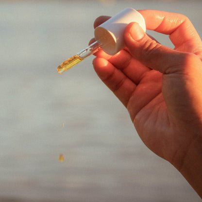 A hand holding the cap off a small white dropper bottle, with a single drop of golden-yellow liquid dripping below. The background is a soft, out-of-focus natural outdoor scene in warm light.