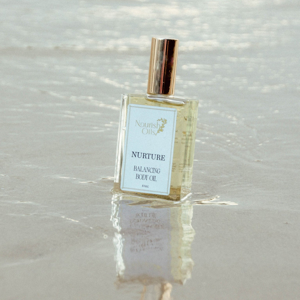 A bottle of North Shore Oils 'Nurture' Balancing Body Oil standing in shallow water on a sandy beach, with its reflection visible on the wet sand.