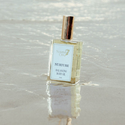 A bottle of North Shore Oils 'Nurture' Balancing Body Oil standing in shallow water on a sandy beach, with its reflection visible on the wet sand.