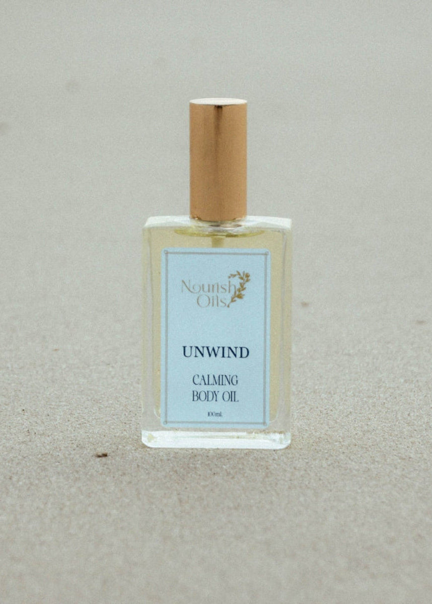 A bottle of Nourish oils 'Unwind Calming Body Oil' with a gold cap and light blue label, placed on a sandy beige background.