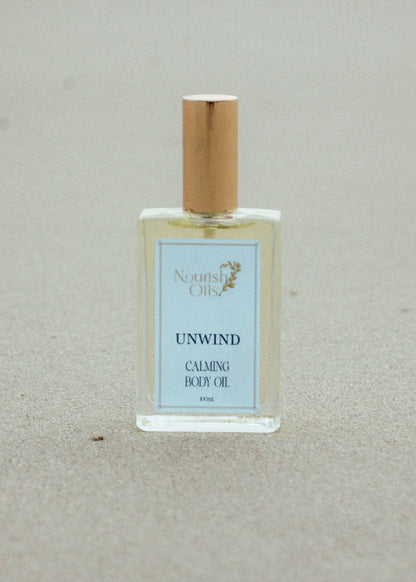 A bottle of Nourish oils 'Unwind Calming Body Oil' with a gold cap and light blue label, placed on a sandy beige background.