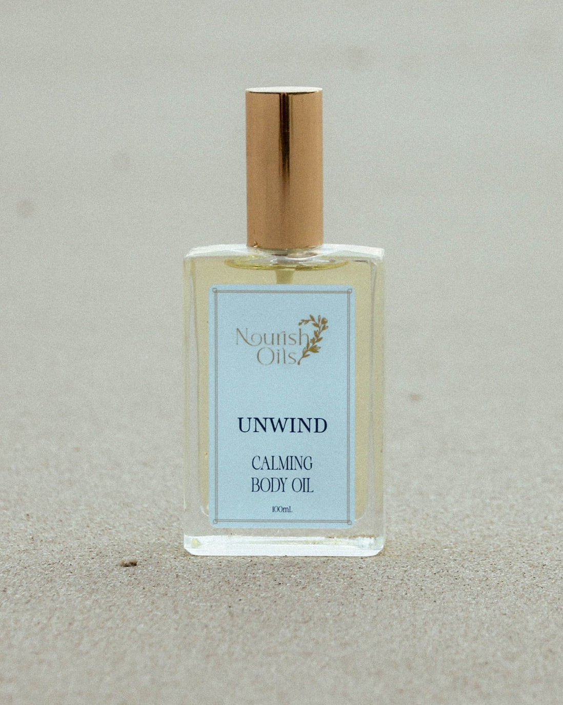 A clear rectangular bottle of North Shore Unwind Calming Body Oil with a gold cap, standing on a sandy beach background.