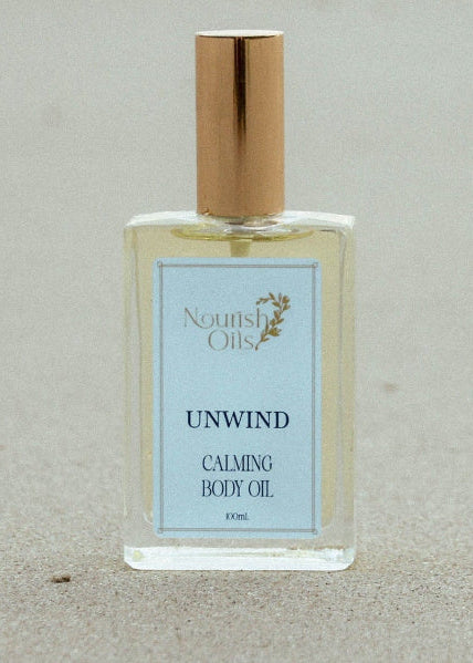 A clear rectangular bottle of North Shore Unwind Calming Body Oil with a gold cap, standing on a sandy beach background.