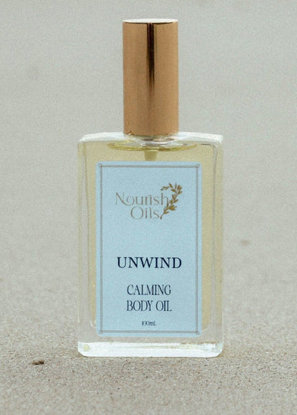 A clear rectangular bottle of North Shore Unwind Calming Body Oil with a gold cap, standing on a sandy beach background.